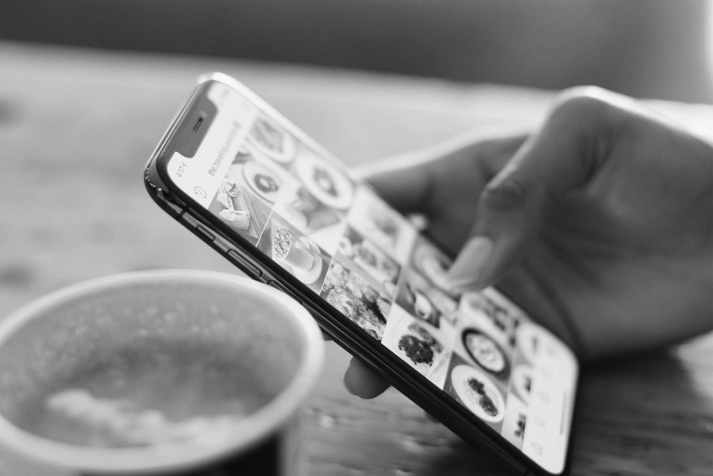 A woman's hand scrolling through social media next to a delicious looking cup of coffee. The image is black and white.