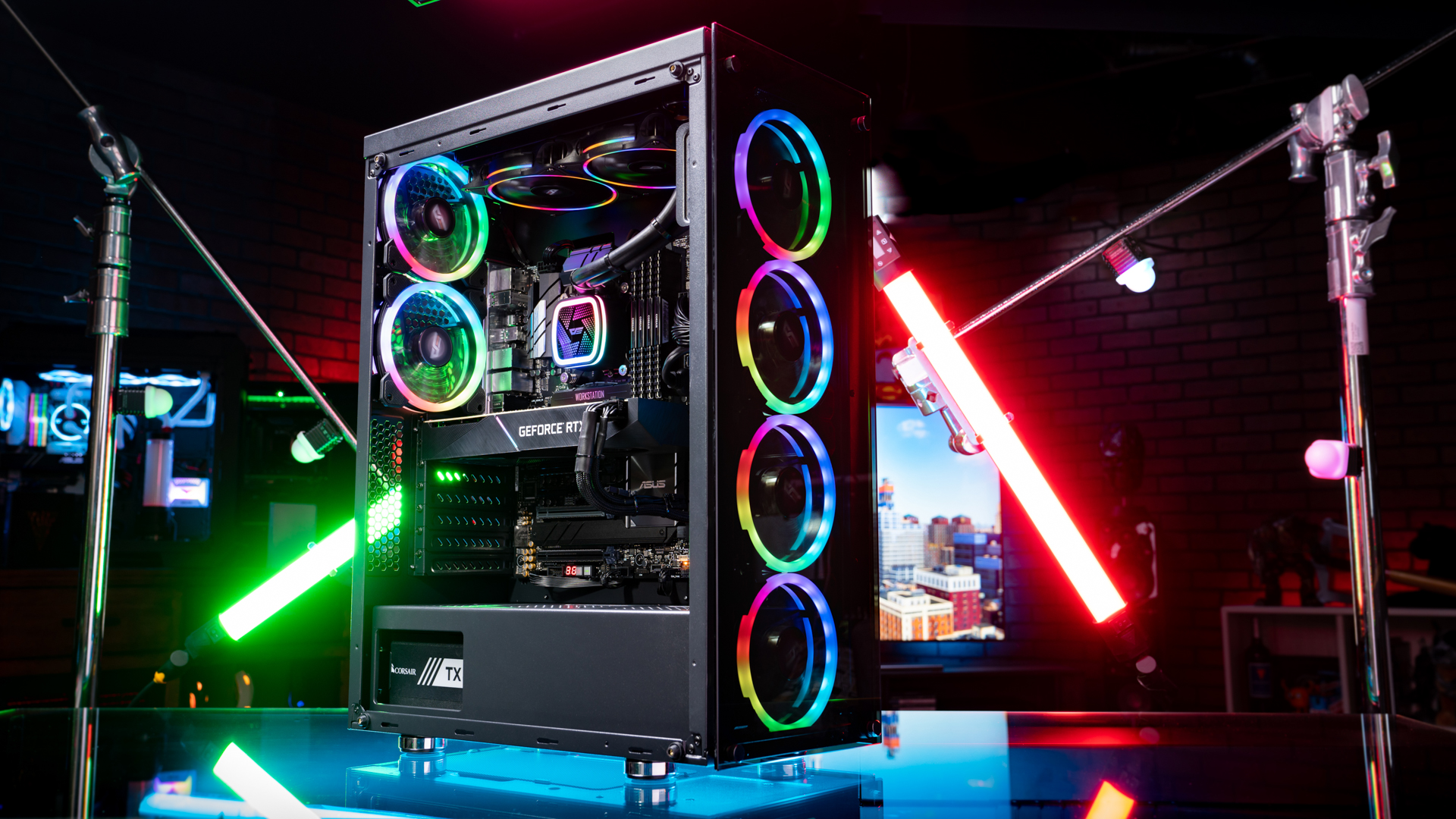 A colorful PC build with flashing RGB lights across the CPU cooler, fans, and RAM. The PC is on a glass table surrounded by bright lights. 