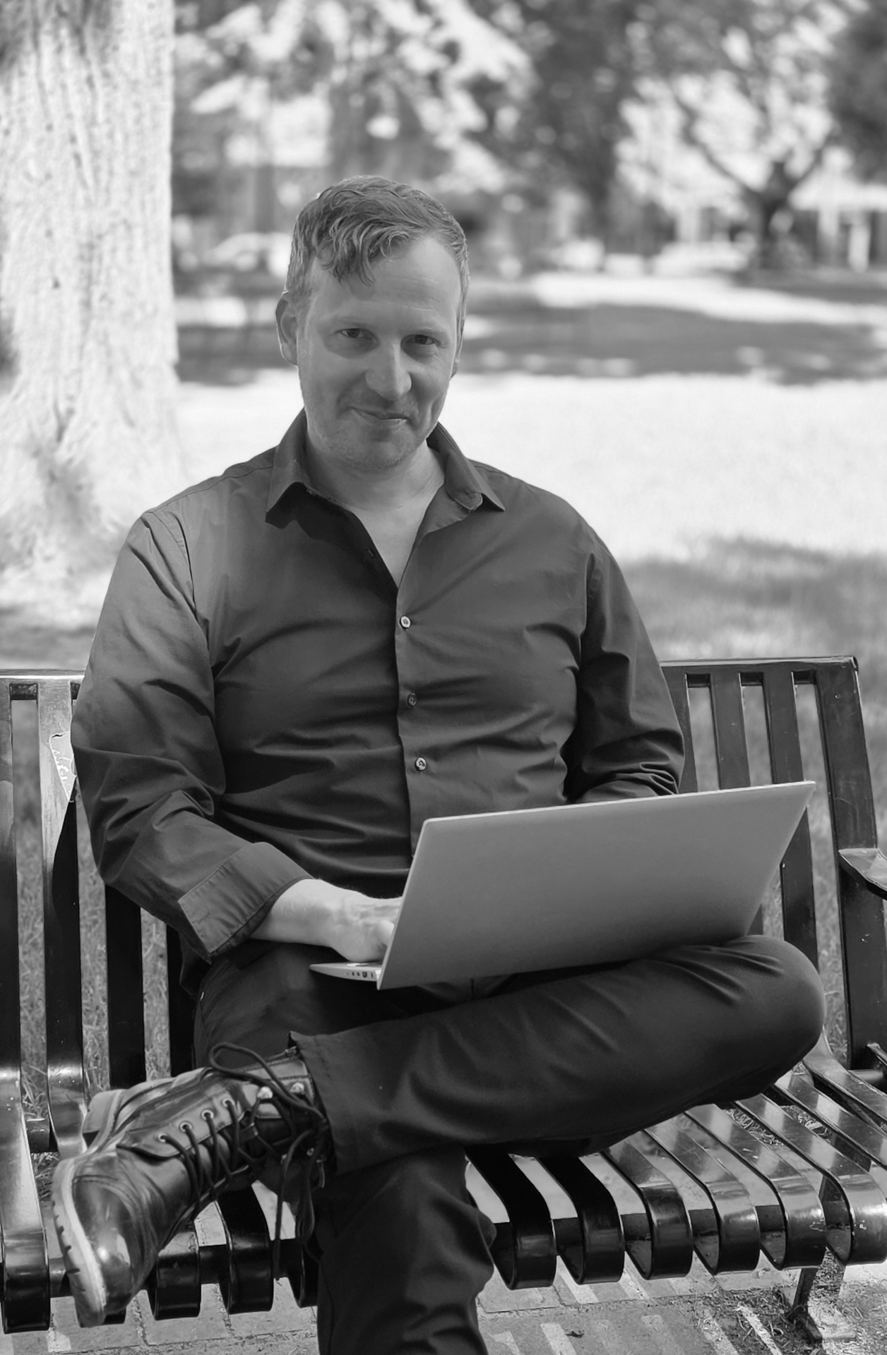 Black and white photo of copywriter Leo Parrill sitting on a park bench with a laptop, looking at the camera. He looks like a fun, confident, and capable professional.